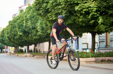 Giysi ve koruyucu kask kamera binicilik bisiklet yeşil ağaçlarıyla çevrili boş şehir merkezi sokak aşağı bakarak Bisiklet sportif adam motorcu. Sporcu eğitim açık havada becerilerini geliştirmek.