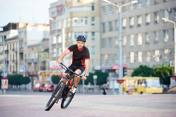 Handsome guy bicyclist posing in cycling clothes and helmet on bike for sport campaign in front of city building. Sportsman exercising outdoors, rest after working day. Concept of healthy lifestyle