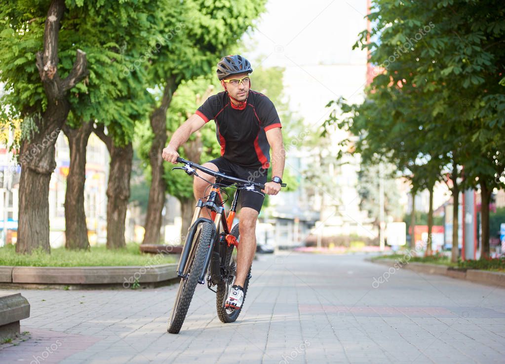 Man professional biker riding on bicycle along empty city streets, green trees around. Sportsman performing morning outdoor exercise and training.