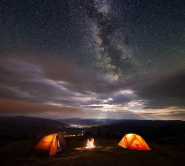 Camping illuminated orange tents on the top of mountain at night under starry sky and milky way on the background of the luminous town which is in the valley. clipart