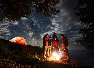 Ağaçlar ve yıldızlar ve Samanyolu ile bulutlu gökyüzü altında ışıklı turuncu çadır kamp ateşi etrafında duran genç turist grubu. Gece dağda kamp