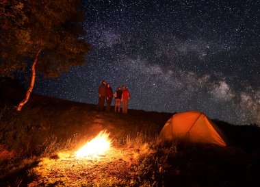 Gece kamp sırasında parlak yıldız ile mayınları alışılmadık gökyüzü zevk yürüyüşçü grubudur. Ön planda, çadır ve ağaçlar bir ateş yakar.