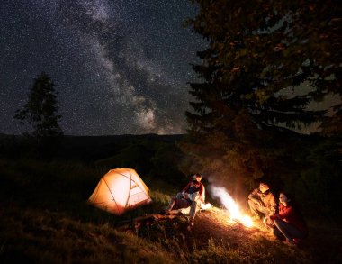 İki yıldızlı gökyüzü Samanyolu ile zevk kamp ateşi, çiftler. Her diğer, diğer tarafta ikinci oturma sarılma günlük turuncu çadır, yanında oturan iki tane. Dağlarda forest yakınındaki kampa gece