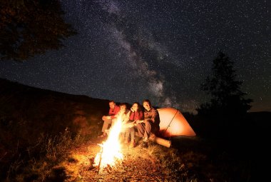 Bir gece yıldızlar ve Samanyolu ile mayınları inanılmaz güzel gökyüzüne karşı dağlarda kamp sırasında bir günlük parlak kamp ateşi yakınındaki bir dinlenme sahip dört mutlu yürüyüşçü.