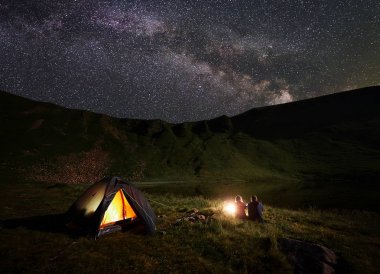 Gece yıldızlar ve parlak Samanyolu ile mayınları gökyüzü altında göl yakınındaki dağlarda kamp. Arkadan görünüş manzaranın tadını bir dinlenme yakınında kamp ateşi ve ışıklı çadır, erkek ve kız yürüyüşçü