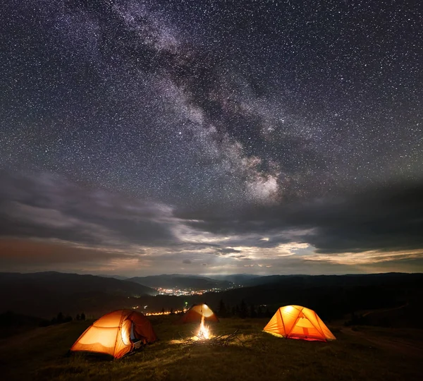 Night camping. Three illuminated orange tents in the mountains at night under starry sky, Milky way and clouds on the background of the luminous town in the valley.