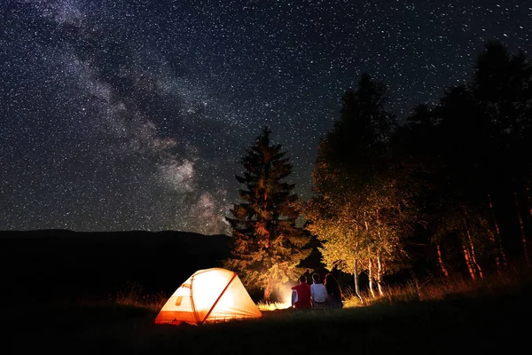 Friends hikers sitting by the campfire at night near the forest and illuminated orange tent under the sky with a huge number of bright stars and milky way on the background of mountains and hills.