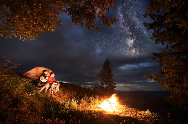 Camping at night. Young couple hikers sitting by the campfire and illuminated orange tent, looking into the distance under the evening sky. Through clouds on the sky are visible bright stars.