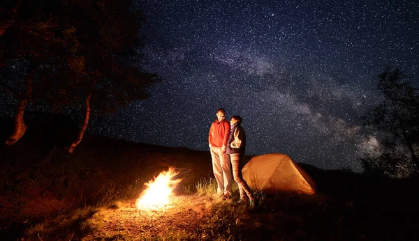 Young couple man and woman hikers smiling to each other, standing near campfire and orange tent during night camping under starry sky and Milky way.