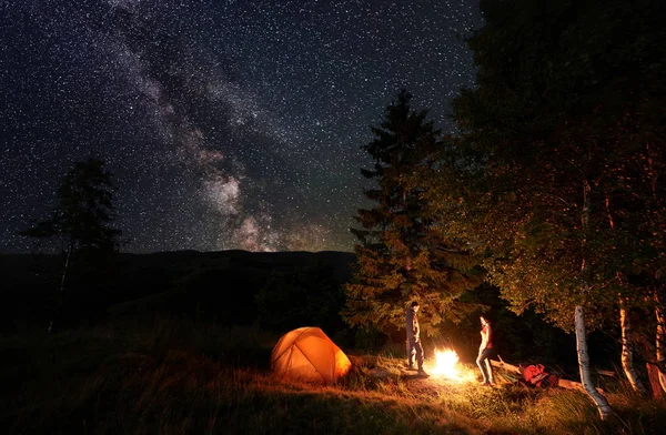 Guy Girl Backpackers Standing Campfire Woods Illuminated Orange Tent Starry — Stock Photo, Image
