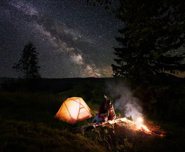 Man pointing to the incredible beautiful starry sky and Milky way. Couple sitting near the glowing tent, trees and bonfire at log in mountains. Romantic evening camping under the fabulous starry sky