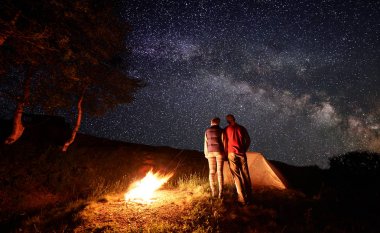 Yıldızlı gökyüzüne bakarak ile Samanyolu, kamp ateşi, çadır ve ağaçlar birbirine yapışan iki kişi backpackers görünümünü arka. Gece dağda kamp, aktif dinlenme kavramı