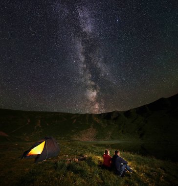Dağ ve göl inanılmaz güzel yıldızlı gökyüzü altında gece kamp sırasında hayran yakınında çadır, kamp ateşi etrafında oturan arka görünümü genç iki kişi yürüyüş yapanlar. İçinde açıkça görünür Samanyolu gökyüzü.