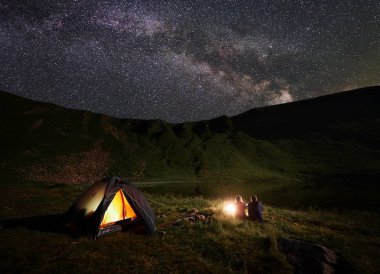 Gece yıldızlar ve parlak Samanyolu ile mayınları gökyüzü altında göl yakınındaki dağlarda kamp. Arkadan görünüş manzaranın tadını bir dinlenme yakınında kamp ateşi ve ışıklı çadır, erkek ve kız yürüyüşçü