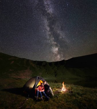 Genç çift backpackers bir dinlenme ışıklı çadırda kamp ateşi, kamp, inanılmaz yıldızlı gökyüzü parlak Samanyolu ile zevk gece sahip kucaklayan. Dağ ve göl arka plan üzerinde