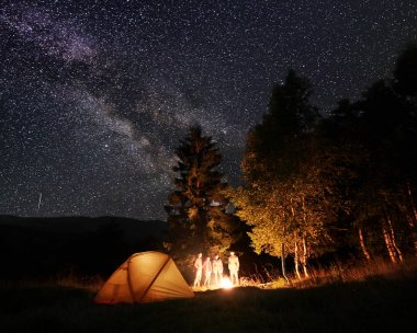 Arka planda dağlar ve tepeler inanılmaz güzel yıldızlı gökyüzü ve Samanyolu altında ateş yanan bakarak ışıklı çadır, forest yakınındaki gece kamp, turist grup.
