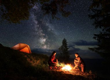 Romantik erkek ve kadın turist kendilerini geceleri ateş etrafında ısınma. Ağaçlar ve ışıklı turuncu çadır yakınındaki dağlarda kamp. Üstlerindeki yıldızlı gökyüzü ve Samanyolu.