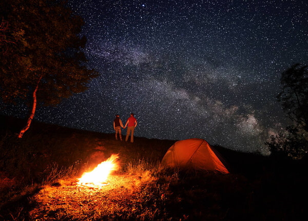 Night camping with a campfire and tent under a bright starry sky and Milky way. Man and woman tourists are holding hands and looking at each other under the romantic sky in the mountains
