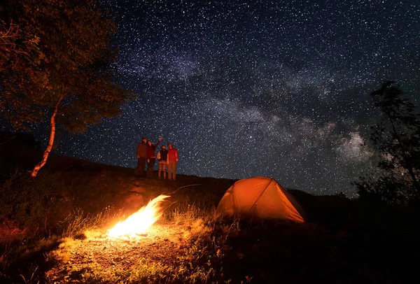 Four person enjoying the unusual sky strewn with bright stars during the night camping. Girl shows the rest of the group up on Milky way. In the foreground, campfire burning near the tent and trees.