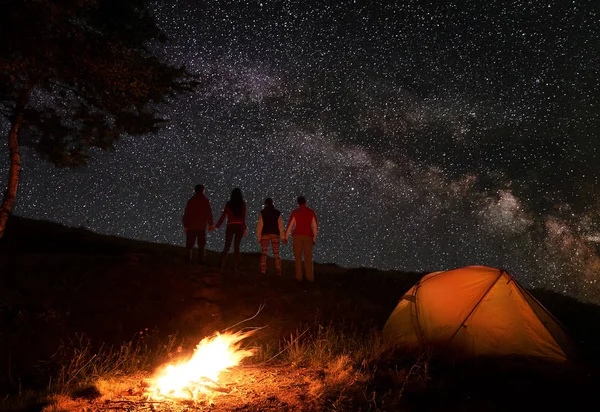 Back view of two pair hikers holding hands and enjoying milky way at starry sky during a night of camping near a campfire and orange tent in mountains. Romantic evening under the fabulous starry sky