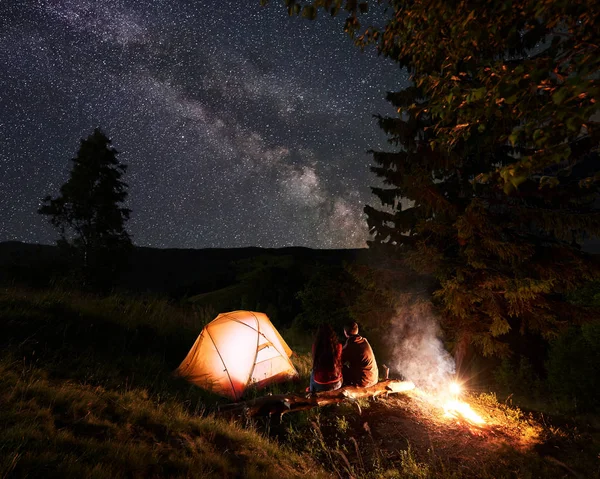 Rear view man and woman sitting on log by the campfire and illuminated orange tent, admiring the scenery of night mountains above which the bright starry sky and milky way. Night camping near forest