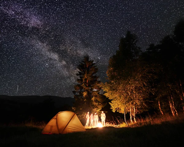 Group of tourists at night camping near the forest at the illuminated tent, looking at the burning fire under incredible beautiful starry sky and Milky way on the background of mountains and hills.