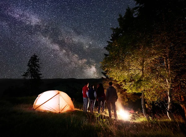Night camping near the forest under a magical starry sky and Milky way on hills background. Young people hikers around the campfire and illuminated orange tent. Rear view. The concept of active rest