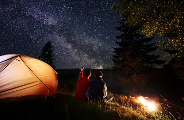 Back view male hiker showing female up on the evening starry sky at Milky way. Couple sitting in the camping near illuminated orange tent, trees and bonfire. Romantic evening under fabulous starry sky