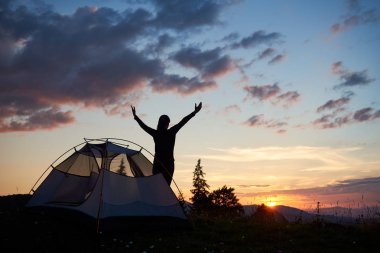 Çadır kır çiçekleri ve köknar ağaçları ile dağın zirvesinde yakınındaki Kollarını açarak şafakta duran görünümü siluet kadın geri. Akşam gökyüzü ve gün batımı dağlar ve tepeler arkasında eşsiz manzarası