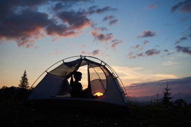 Arka görünümü bir çadırda akşam mavi gökyüzü altında oturan kız Yüce Dağların üzerinden günbatımı parlak güneşin arka plan üzerinde zevk silüeti. Bir çadır görüntülemek