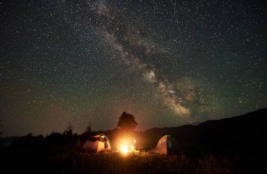 Muhteşem gece gökyüzünde yıldızlar ve Samanyolu dolu altında dağlarda gece kamp istirahat kadın kamp. Kız uzun yürüyüşe çıkan kimse kamp ateşi ve iki çadır yanında oturuyor. Aktif yaşam tarzı kavramı. Astrophotography