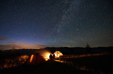 Arka görünümü muhteşem gece gökyüzüne yıldız ve Samanyolu, zevk akşam sahne dolu arayan iki çadır, günlük kamp ateşi, yanında oturan dağda kamp adlı iki oğlu yürüyüşçü ve siluet anne