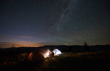 Arkadan görünüş anne ve günlük kamp ateşi ve parlayan çadır, yanında oturan dağlarda gece kamp, bir dinlenme iki çocuk turist güzel yıldızlı gökyüzü ve Samanyolu akşamları bakarak