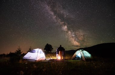 Arkadan görünüşü anne ve kamp ateşi ve iki ışıklı çadırlar, yanında duran Dağları, kamp dinlenme iki oğlu backpackers gece gökyüzüne yıldızlarla, dolu Samanyolu, gece sahne zevk arıyorsunuz.
