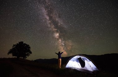 Etkin mutlu kadın backpacker gece dağda kamp, bir dinlenme. Işıklı çadır inanılmaz gece gökyüzünde yıldızlar ve Samanyolu dolu manzarayı elleriyle kaldırdı, yanında kadın ayakta