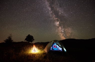Kadın uzun yürüyüşe çıkan kimse gece kamp ateşi altında inanılmaz güzel yıldızlı gökyüzü ve Samanyolu dağlarda yanında kamp, bir dinlenme. Işıklı çadır ve uyku tulumu içinde oturan kız. Astrophotography