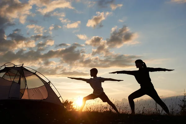 Silhouette view of Yoga Warrior pose exercising by family couple in grass near tent at the daybreak on the background of morning sky with sparse clouds and bright sun. Mom grafts a son\'s love of sport