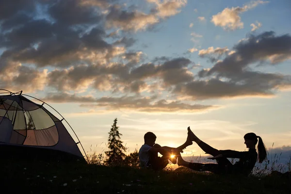 女孩和男孩的剪影坐在草地上的瑜伽姿势在黎明附近的帐篷 女人和男孩的腿在连接 明亮的傍晚的阳光照耀在他们之间的天空 — 图库照片