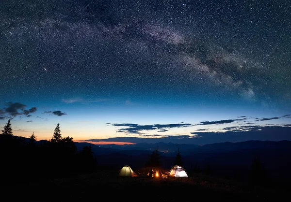 Family tourists mother, father, two sons resting at night camping in mountains, sitting on log beside campfire and two illuminated tents, enjoying amazing view of evening sky full of stars, Milky way