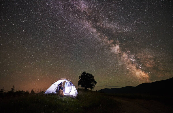 Woman camper enjoying at night camping in the mountains under amazing night sky full of stars and Milky way. Girl sitting inside illuminated tent and looking at sky full of stars. 