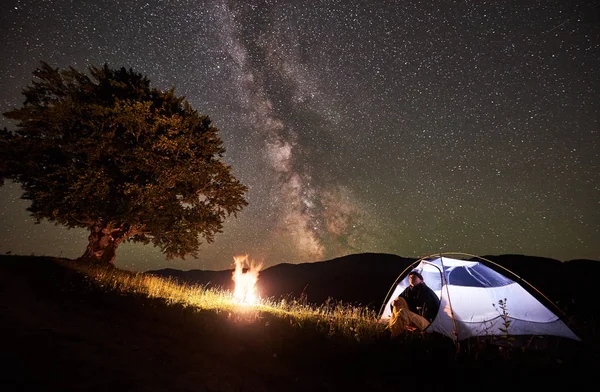 Woman backpacker resting at night camping in the mountains under amazing night sky full of stars and Milky way. Girl hiker sitting inside illuminated tent near campfire and big tree. Astrophotography