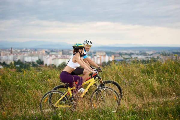Two happy cyclists - man and woman riding on a hill in the grass with wild flowers, city and mountains in the distance.