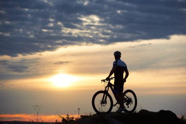 Arkadan görünüşü Bisiklet duran ve şaşırtıcı günbatımı, bulutlar ve gökyüzü harika manzarasına gözlemleyerek sağlam bisikletçi. Kask ve tepede alacakaranlıkta poz spor giyim atletik gizli adam.
