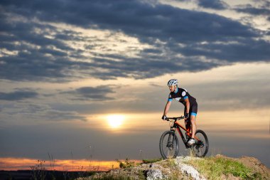 Profesyonel erkek bisikletçi bisiklet dağ Bisiklet rock tepe üzerinde coşku ile. Spor giyim, kask ve spor bardaklarda enerjik ve atletik bir adam. Akşam gökyüzü ve güneş güzel arka plan.