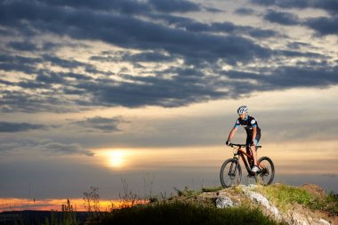 Profesyonel bisikletçi binme dağ bisikleti rock tepe üzerinde coşku ile. Spor giyim, kask ve spor bardaklarda enerjik ve atletik bir adam. Akşam gökyüzü ve güneş güzel arka plan.