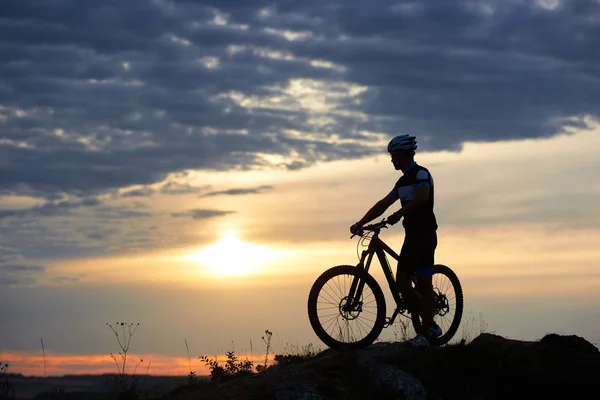 运动的男子在运动衫和头盔手牵着自行车 站在它附近 在山上的黄昏和日落的精力充沛的自行车运动员和天空和云彩的精彩背景 — 图库照片