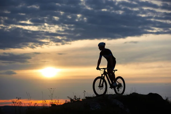 Silhouette of energetic and active cyclist standing on bicycle. Athletic man in helmet and sportswear posing on hill in twilight. Amazing background of sunset and cloudy evening sky.