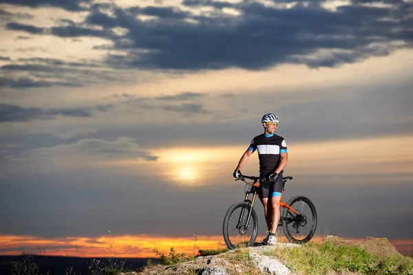 Handsome male cyclist smiling and standing with bicycle on rock hill. Positive sportsman holding his hands on handle bar of bike and posing. Concept of healthy lifestyle, sports and motivation.