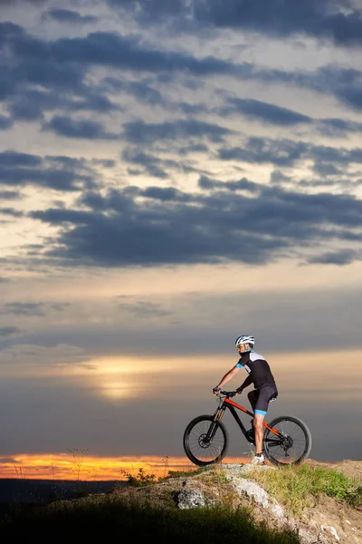 坐在山地自行车上的运动骑自行车的侧面观看 并观察美丽的风景和令人惊叹的日落在多云的天空 男子在黑色运动服和头盔摆在岩石山上 — 图库照片