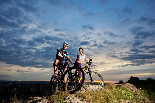 微笑年轻夫妇骑自行车的头盔坐在山地自行车和看相机 — 图库照片